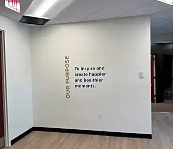 Office hallway with a white wall displaying vertical gold text 'OUR PURPOSE' and blue text 'To inspire and create happier and healthier moments.' Exit sign visible above.